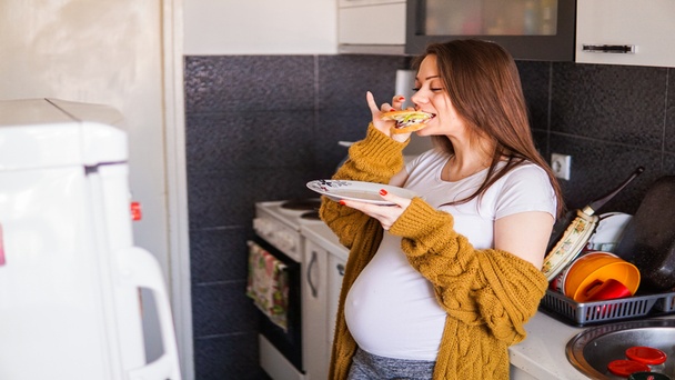 אישה בהריון בשליש השלישי יושבת בבית ואוכלת סנדוויץ' מזין, מייצגת דוגמה למאכלים שכדאי לאכול בהריון בשלב טרימסטר 3 כחלק מתזונה בריאה לאם ולעובר.